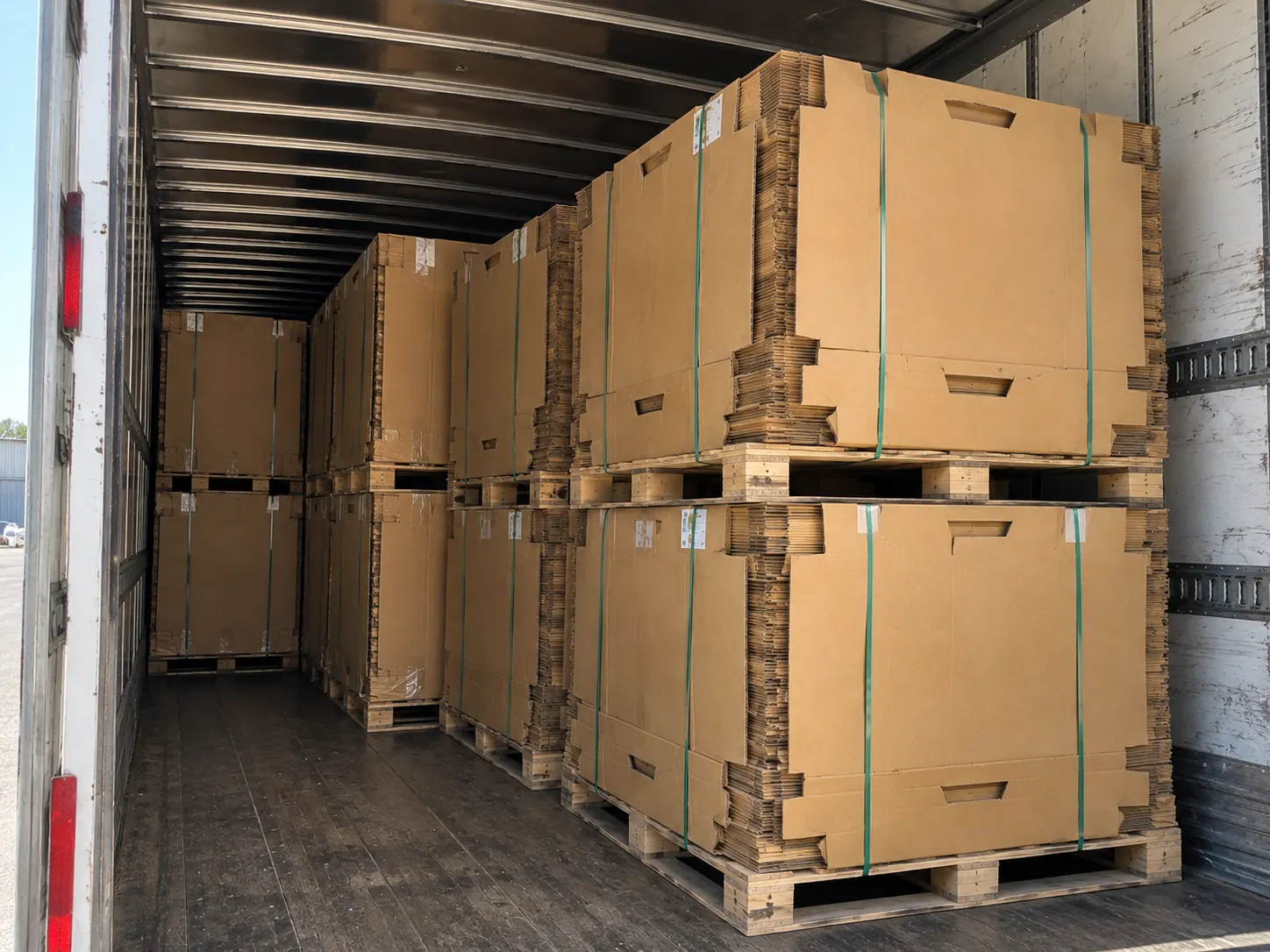 Pallets of knocked-down corrugated board banded flat, stacked in an outdoor yard beside a roller shutter door